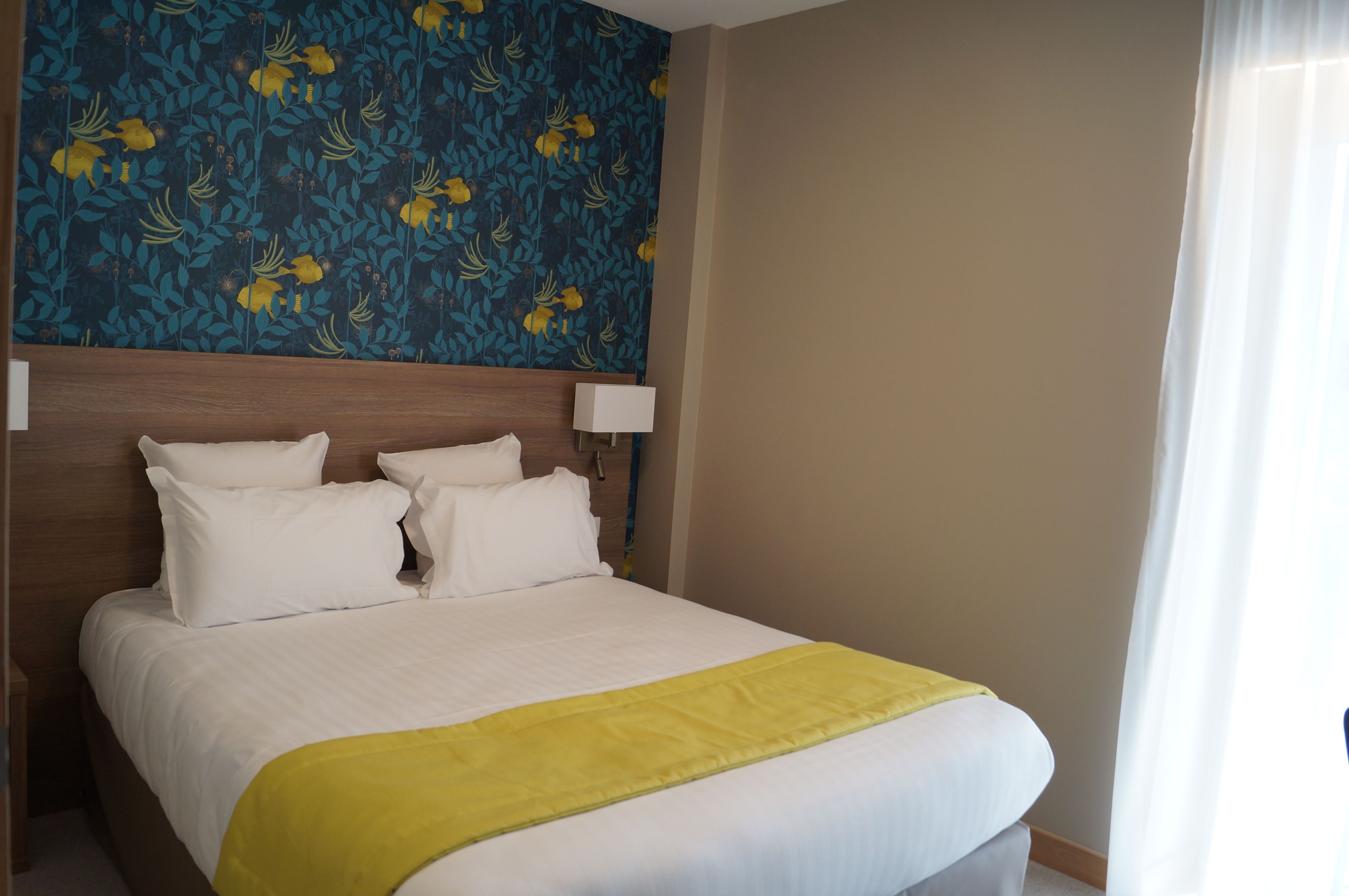 Een net opgemaakt bed met witte lakens en een gele deken, een houten hoofdbord en een wand met een bloemenpatroon.