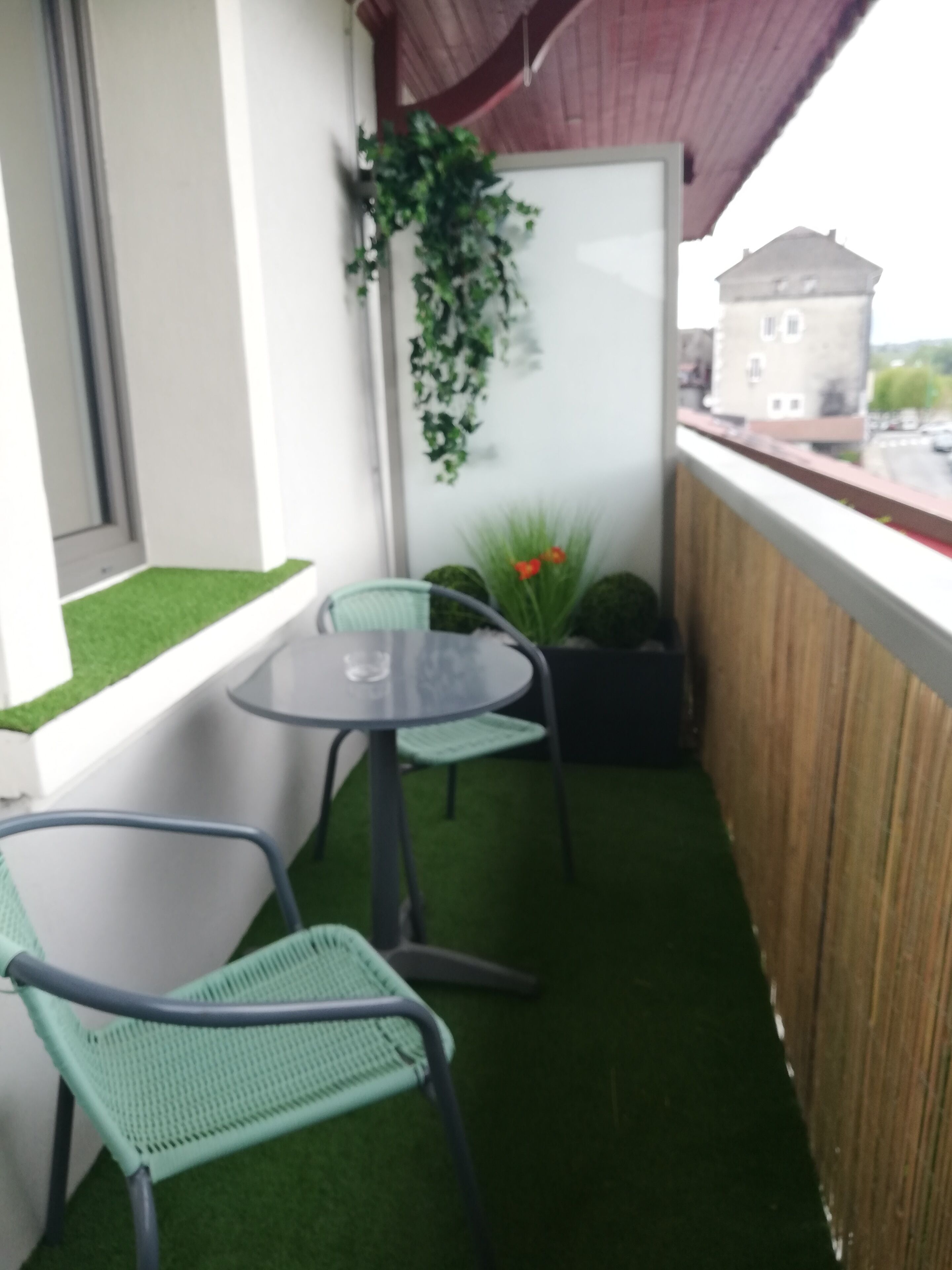 A small balcony with artificial grass, a round table, two green chairs, and a hanging plant.