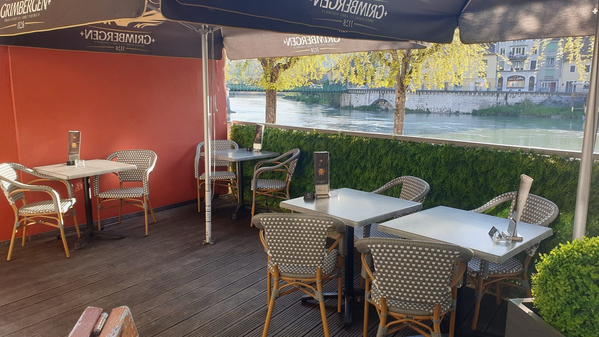 An outdoor seating area with wicker chairs and tables, overlooking a river with a stone bridge in the background.