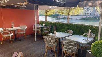 An outdoor seating area with wicker chairs and tables, overlooking a river with a stone bridge in the background.
