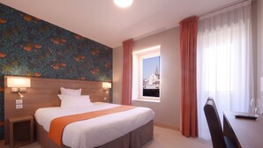 Un cuarto de hotel con una cama grande, una mesita de noche con lámpara, un ventanal con vista a un edificio y un escritorio con silla.
