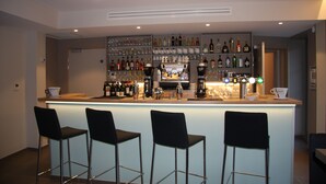 Una zona de bar con mostrador, varios estantes llenos de botellas y cuatro taburetes negros.