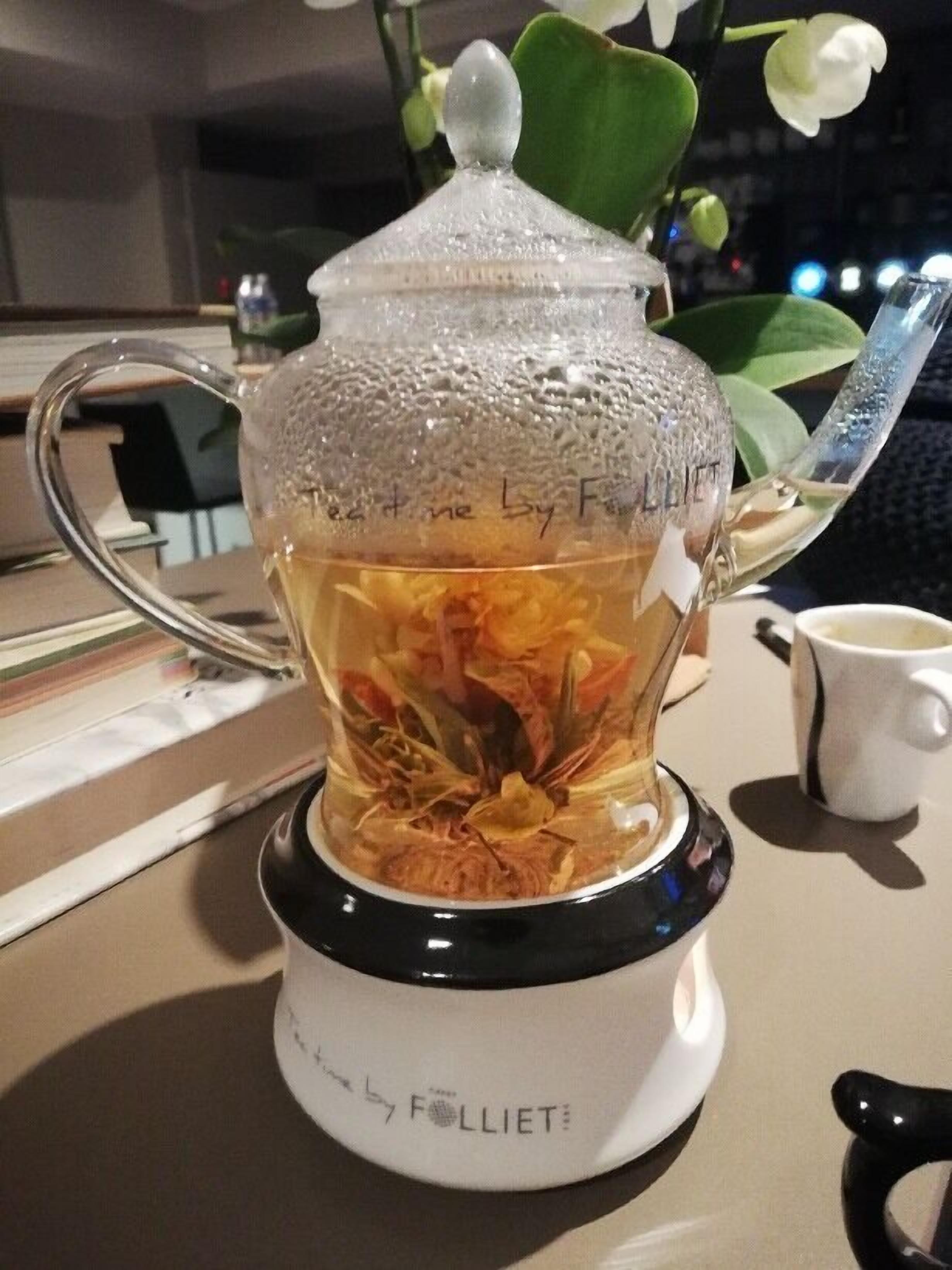 A tea infuser with a transparent lid and a handle, placed on a table with a white mug beside it.