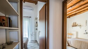 Ein schmaler Flur führt zu einem Schlafzimmer mit einem Bett, einem kleinen Tisch und einem Stuhl. Regale beherbergen Bücher und Dekorationsobjekte.