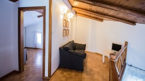 Ein gemütliches Zimmer mit Holzbalkendecke, einer Couch, einem kleinen Tisch mit Fernseher und einem Fenster mit Vorhängen.