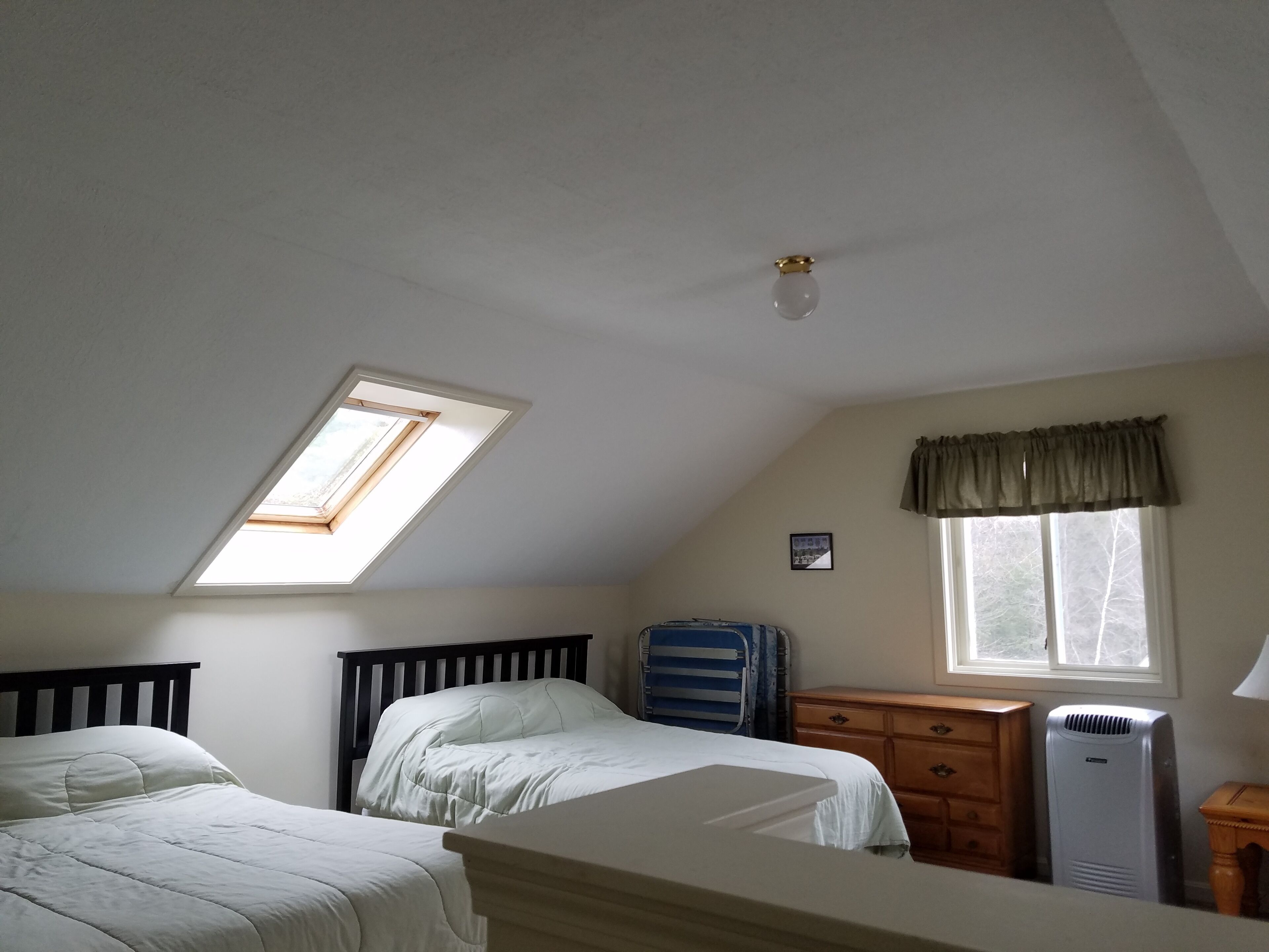A bedroom with two beds, a dresser, a radiator, a window with curtains, and a skylight.