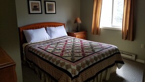 A bedroom with a wooden bed, two framed pictures on the wall, a nightstand with a lamp, and a window with curtains.