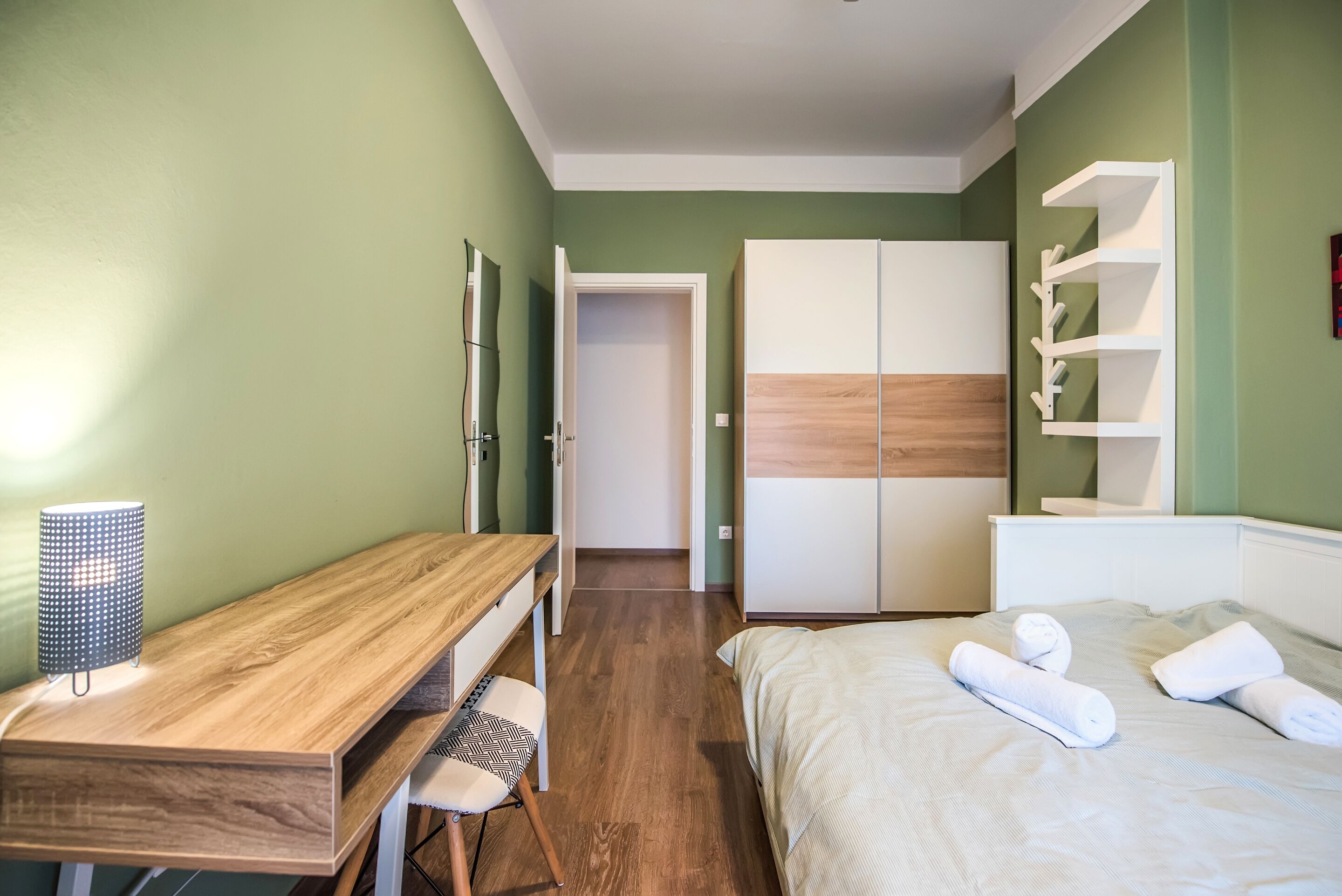 Un dormitorio con un escritorio de madera, una cama con ropa de cama blanca, un armario y una estantería.