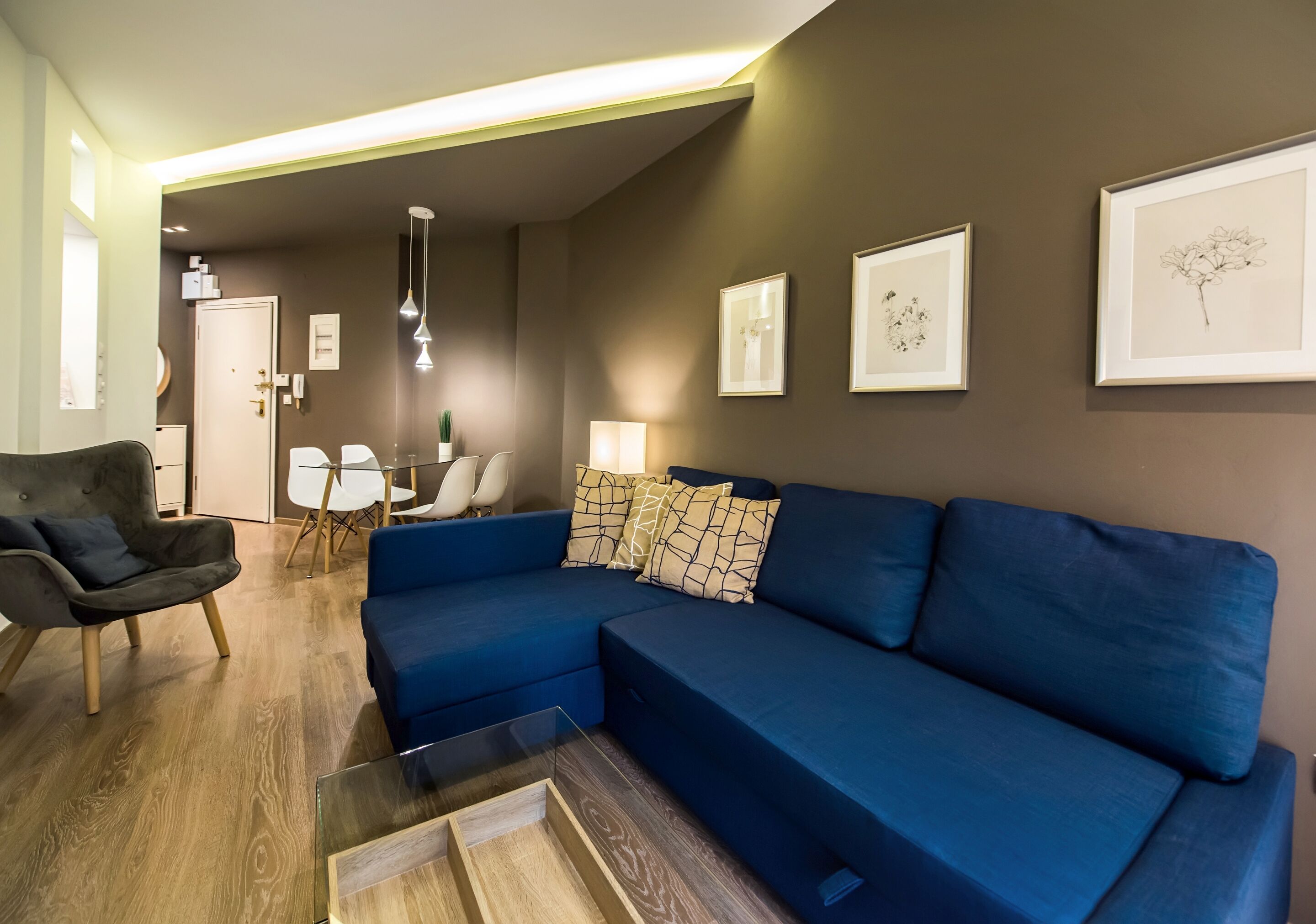 A modern living room with a blue sectional sofa, a dark armchair, and a glass coffee table. There are framed artworks on the walls and a dining area in the background.