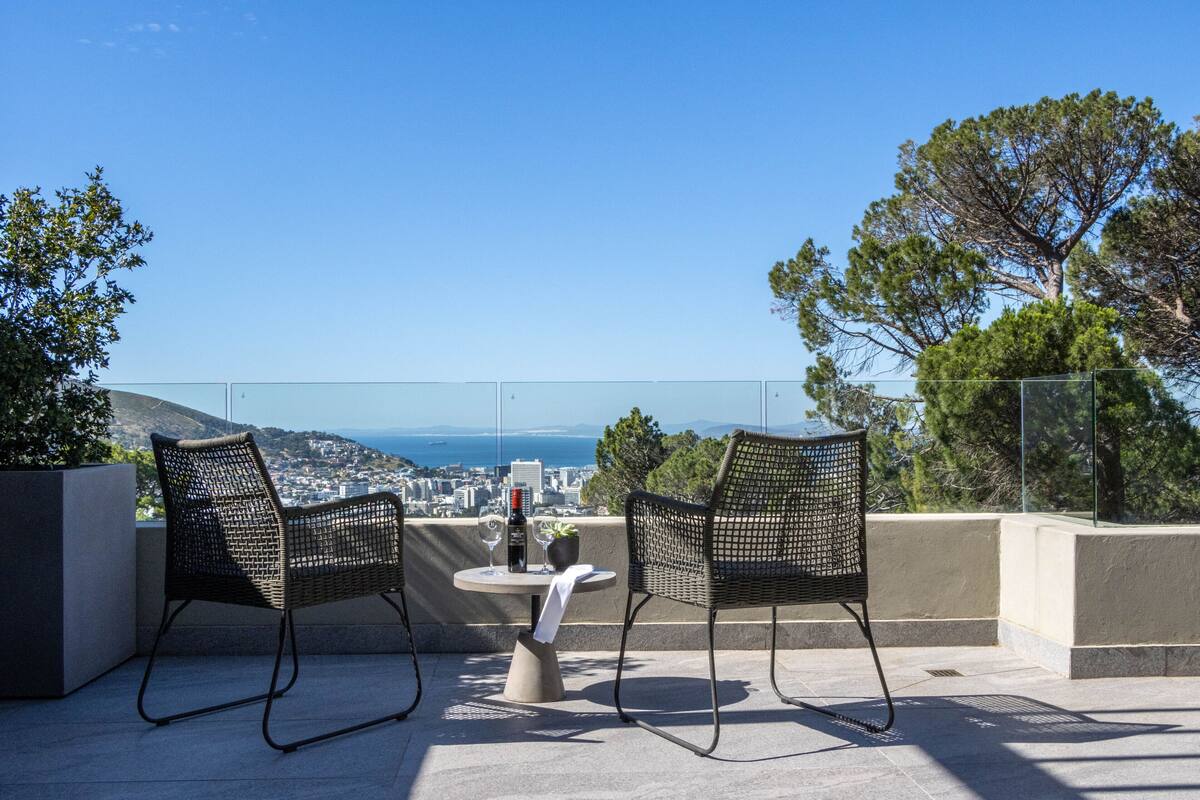 Two wicker chairs and a table with a bottle and glass on a terrace overlooking a city and the sea.