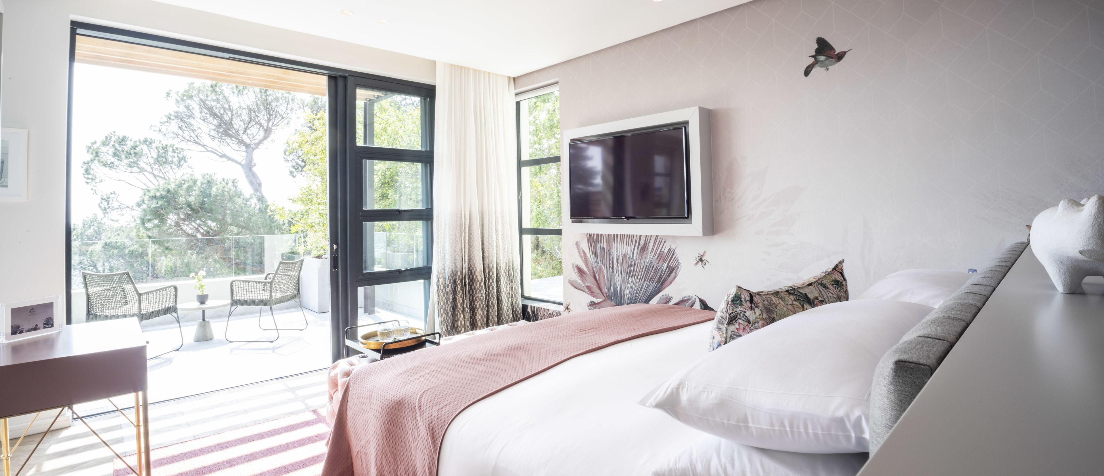 A bedroom with a bed, a TV, a balcony with chairs, and a view of trees.