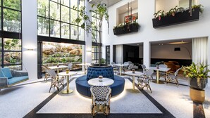 A spacious indoor area with multiple seating arrangements, including round tables and chairs, a central seating area with a blue cushioned bench, and a view of an outdoor garden through large windows. - Camissa House (Cape Town)