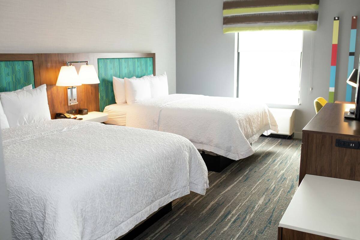 A hotel room with two beds, a desk, a lamp, and a window with a view.