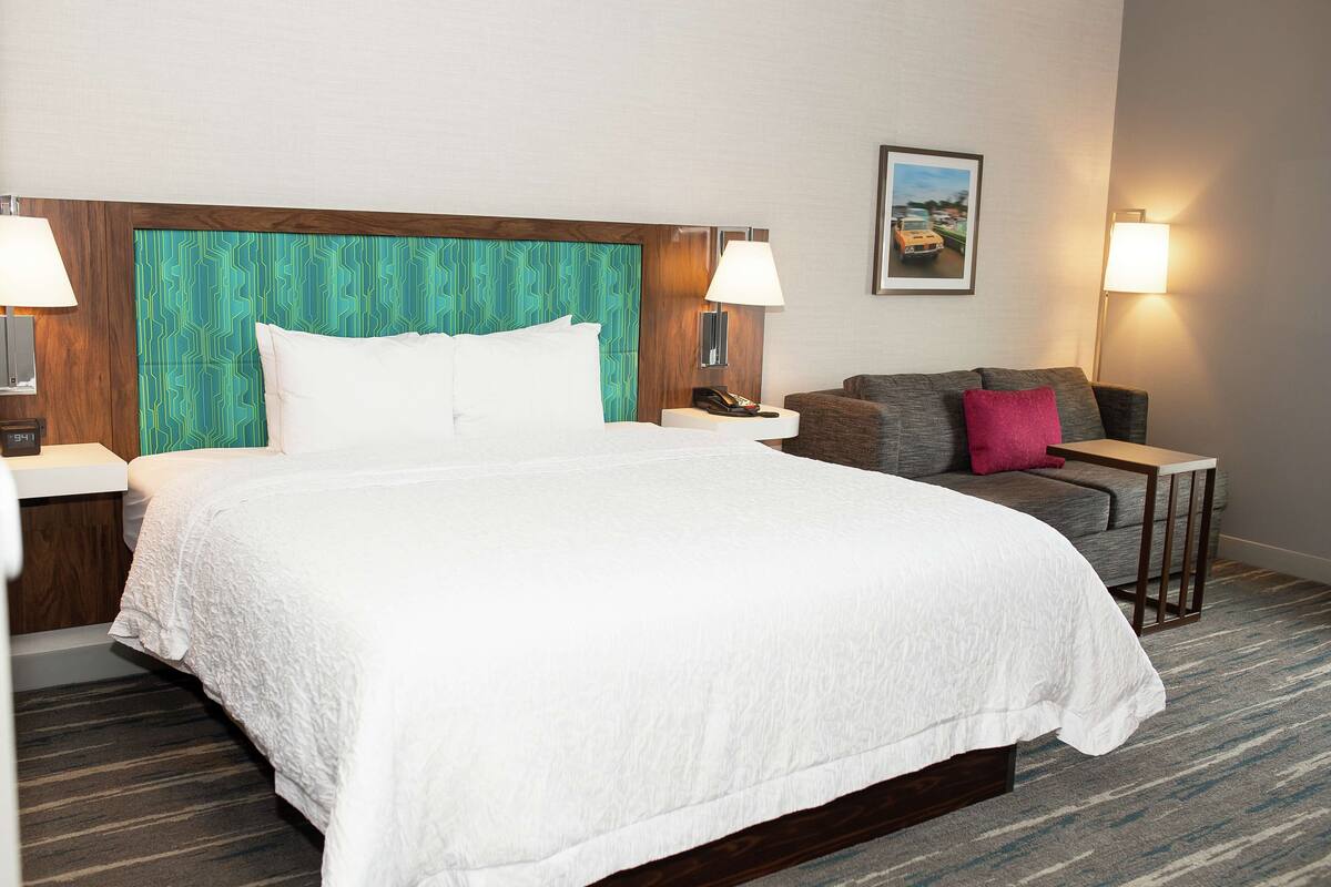 A hotel room with a large bed, a grey sofa, a small table, and two lamps.