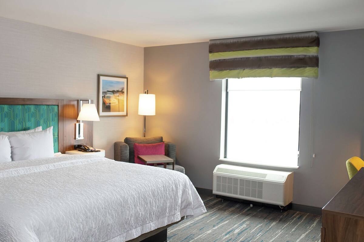 A hotel room with a bed, a desk, a chair, a lamp, a picture on the wall, and a window with a view.