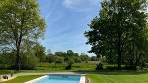 Una piscina rectangular con borde de piedra, rodeada por un césped cuidado y árboles.