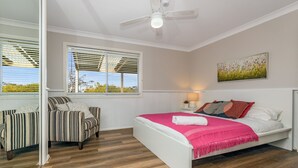 Une chambre à coucher comprenant un lit, une chaise, une fenêtre avec des stores, un ventilateur de plafond et un tableau accroché au mur.