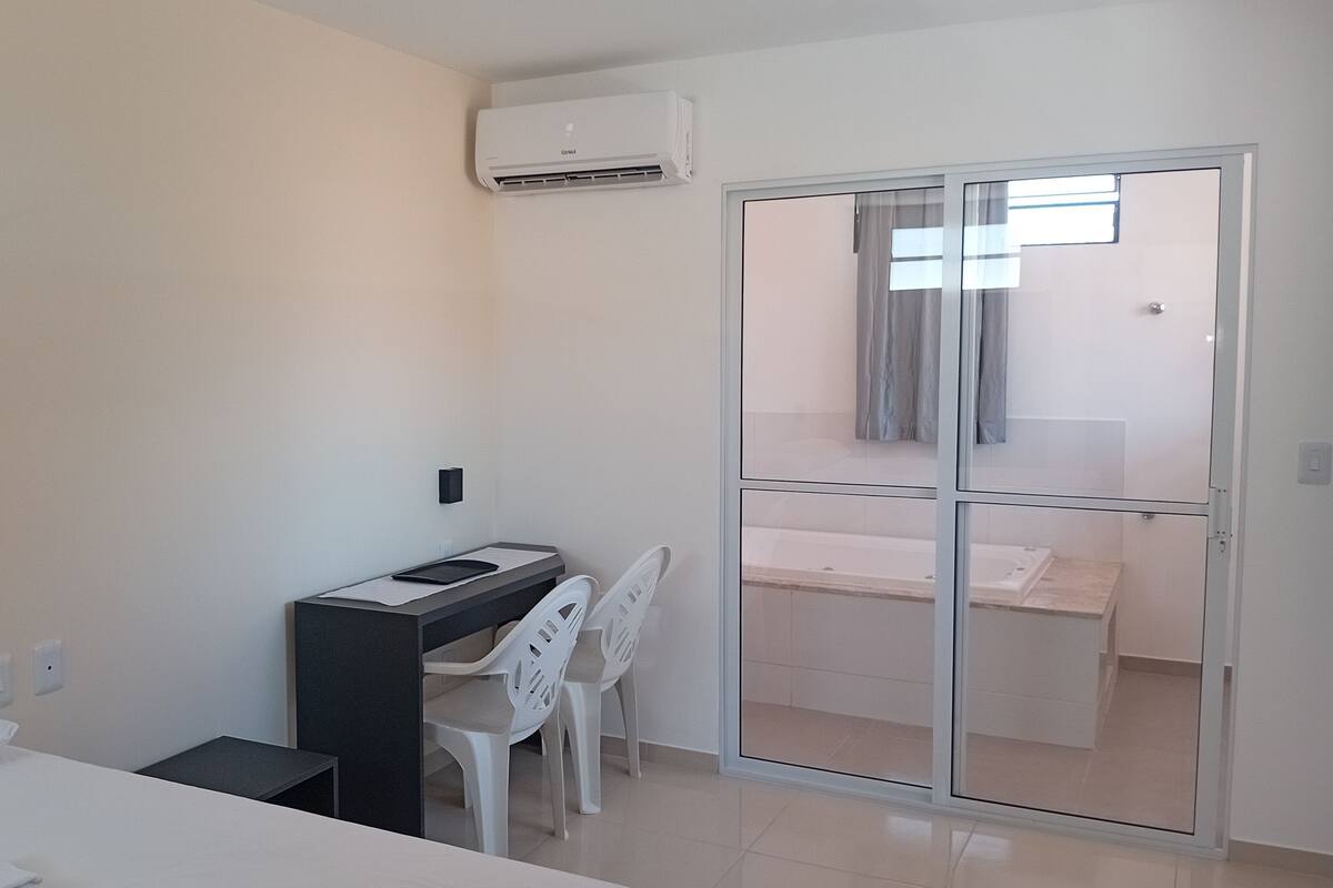A bedroom with a bed, a desk with a chair, an air conditioning unit, and a sliding glass door leading to a bathroom.