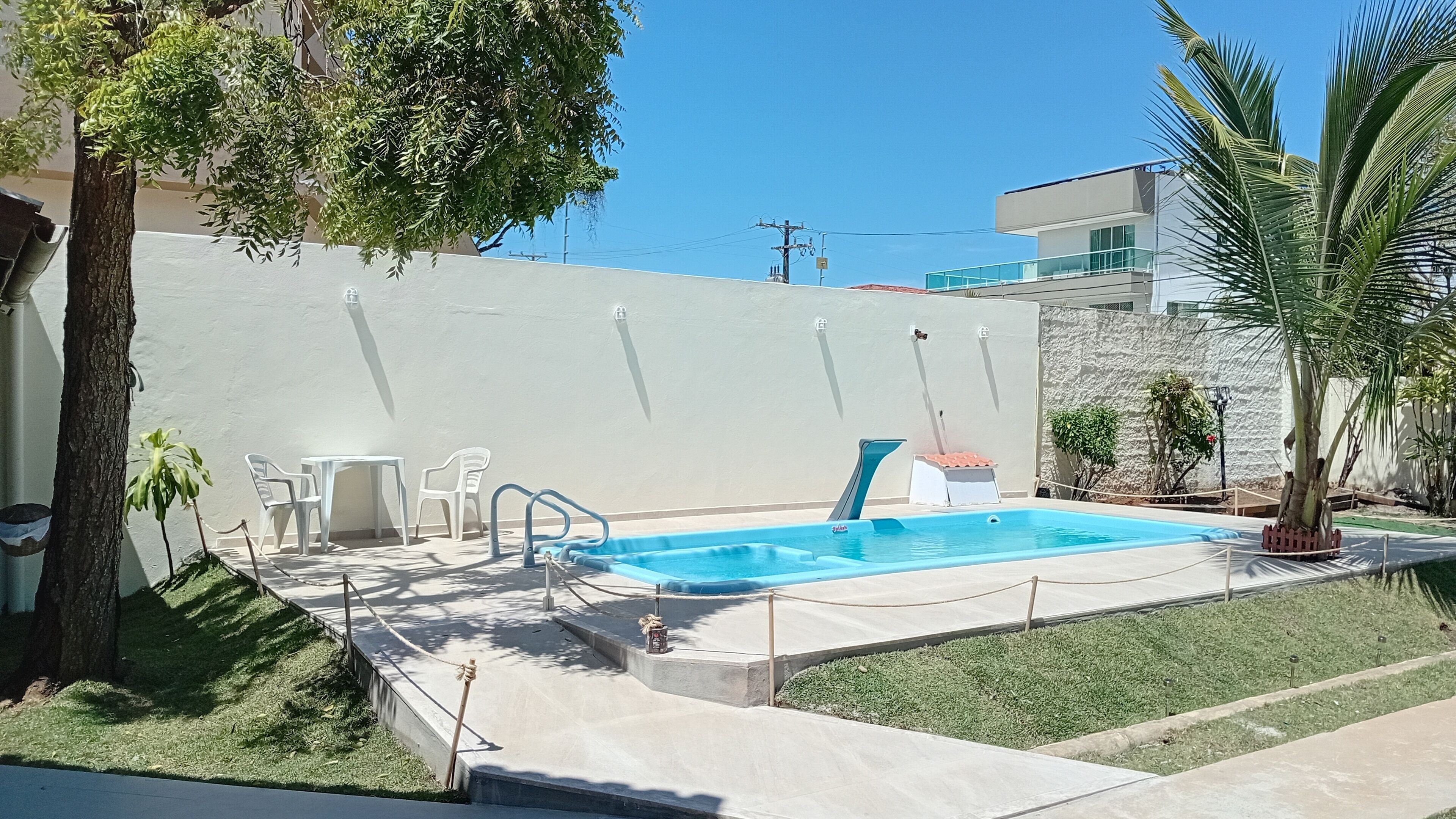 A swimming pool with a small deck area, surrounded by a white wall and a grassy lawn.