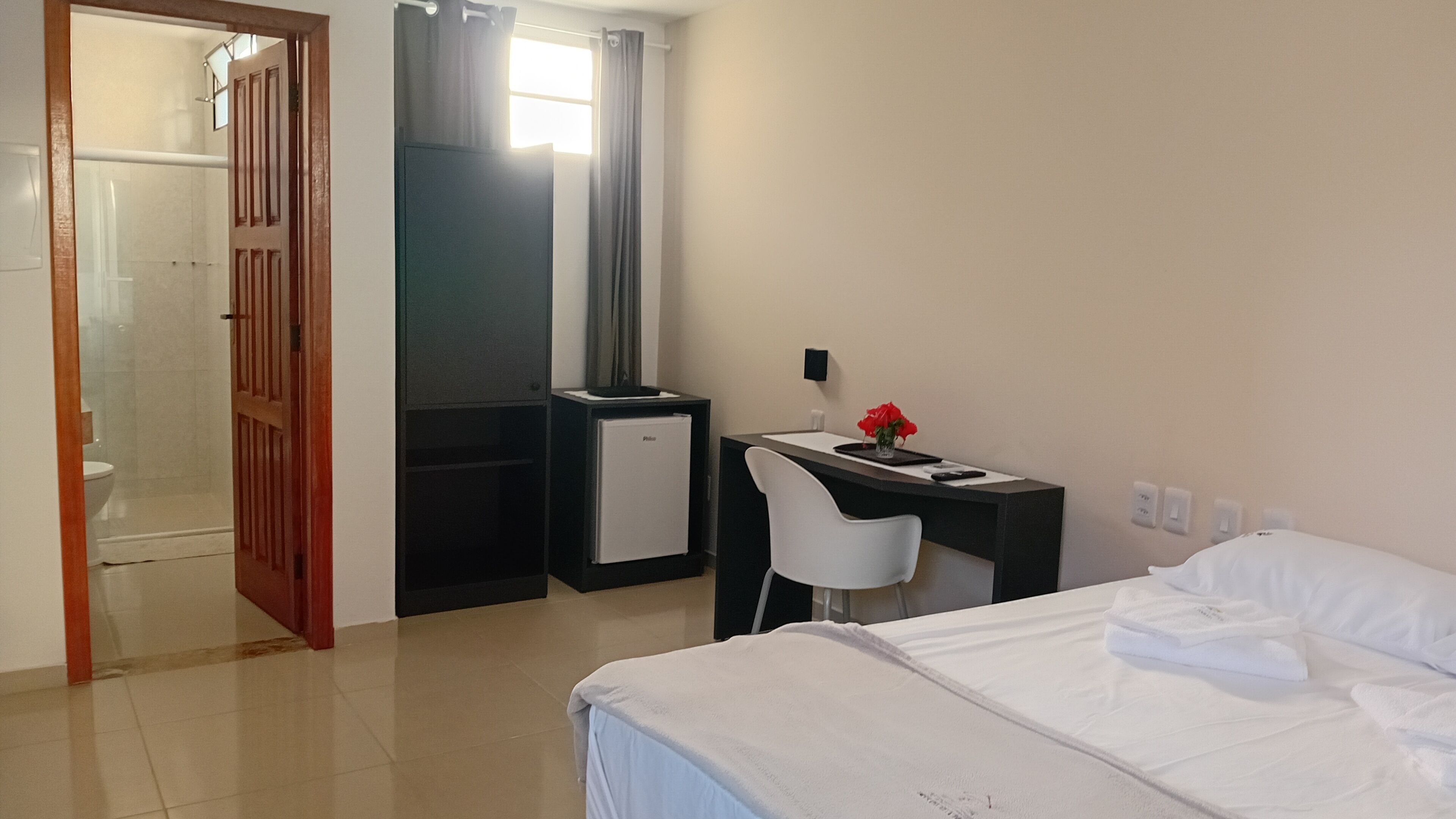 A hotel room with a bed, a desk with a chair, a small refrigerator, and a shower area.