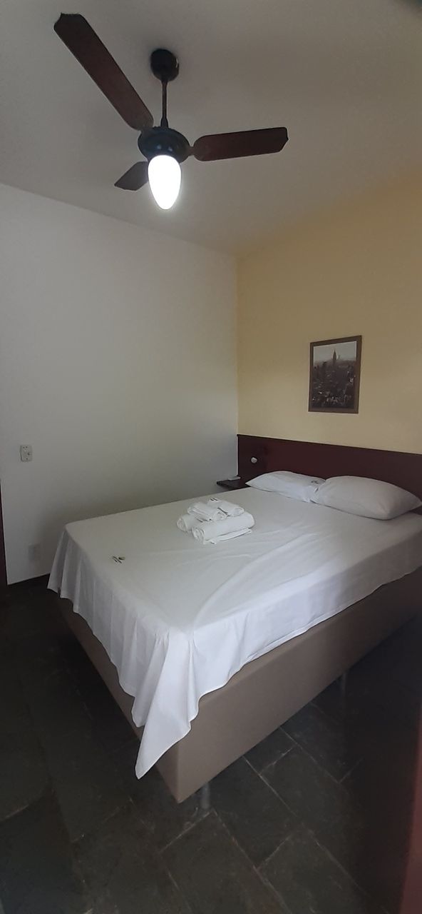 Una cama individual con una colcha blanca y una toalla doblada con cuidado, un ventilador de techo y un cuadro en la pared.