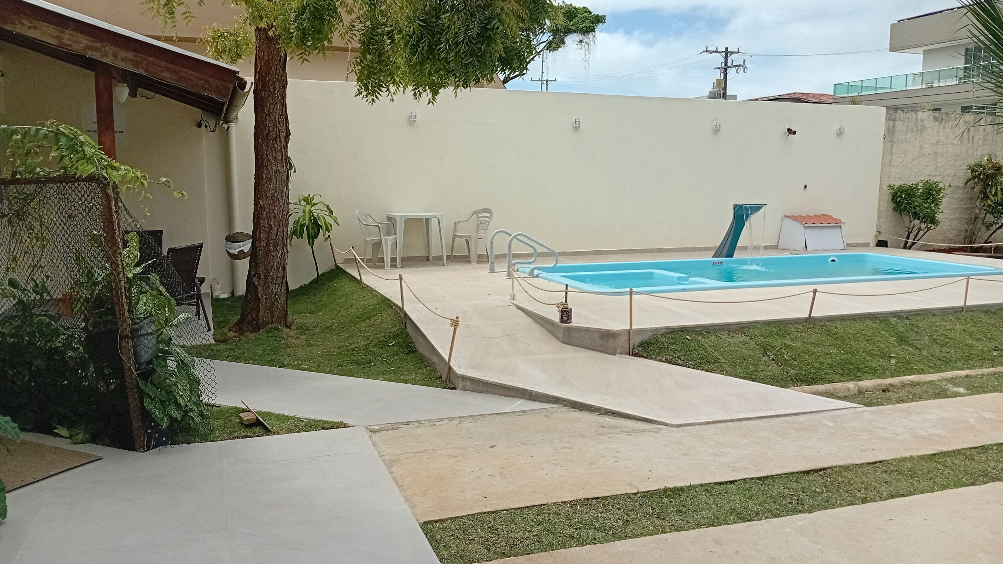 A swimming pool with a small waterfall feature, surrounded by a concrete deck and a grassy area. There are white plastic chairs and a table nearby.