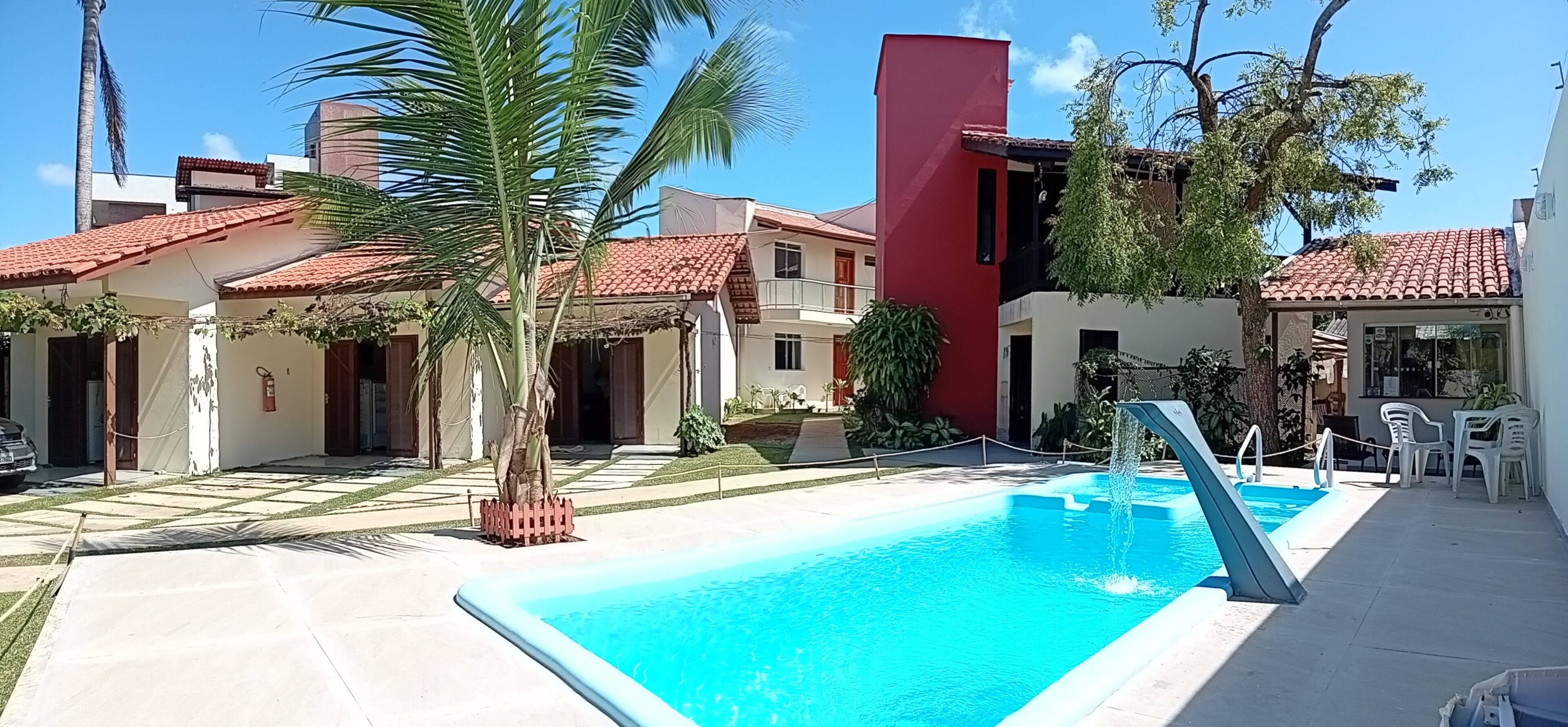 A swimming pool with a water fountain, surrounded by buildings with red-tiled roofs, palm trees, and a clear blue sky.