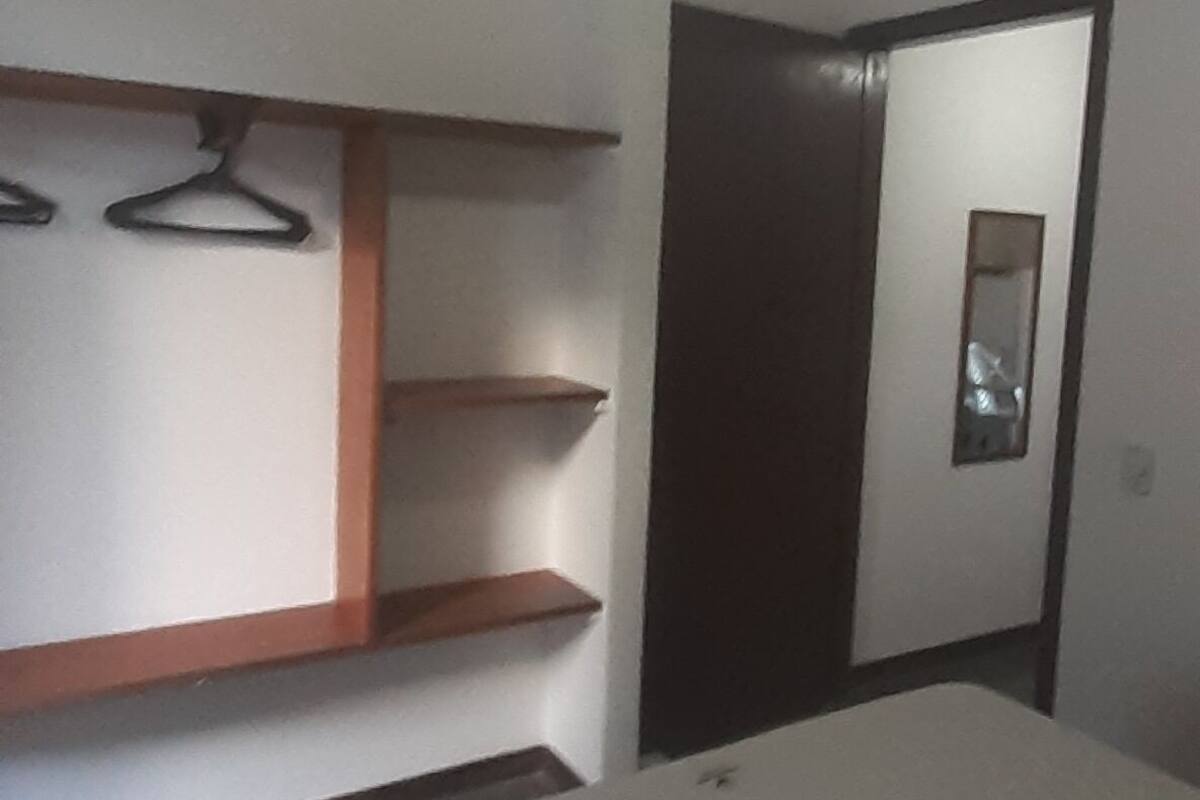 A hotel room with a bed, a ceiling fan, a wall-mounted shelf with hangers, and a door leading to another room.