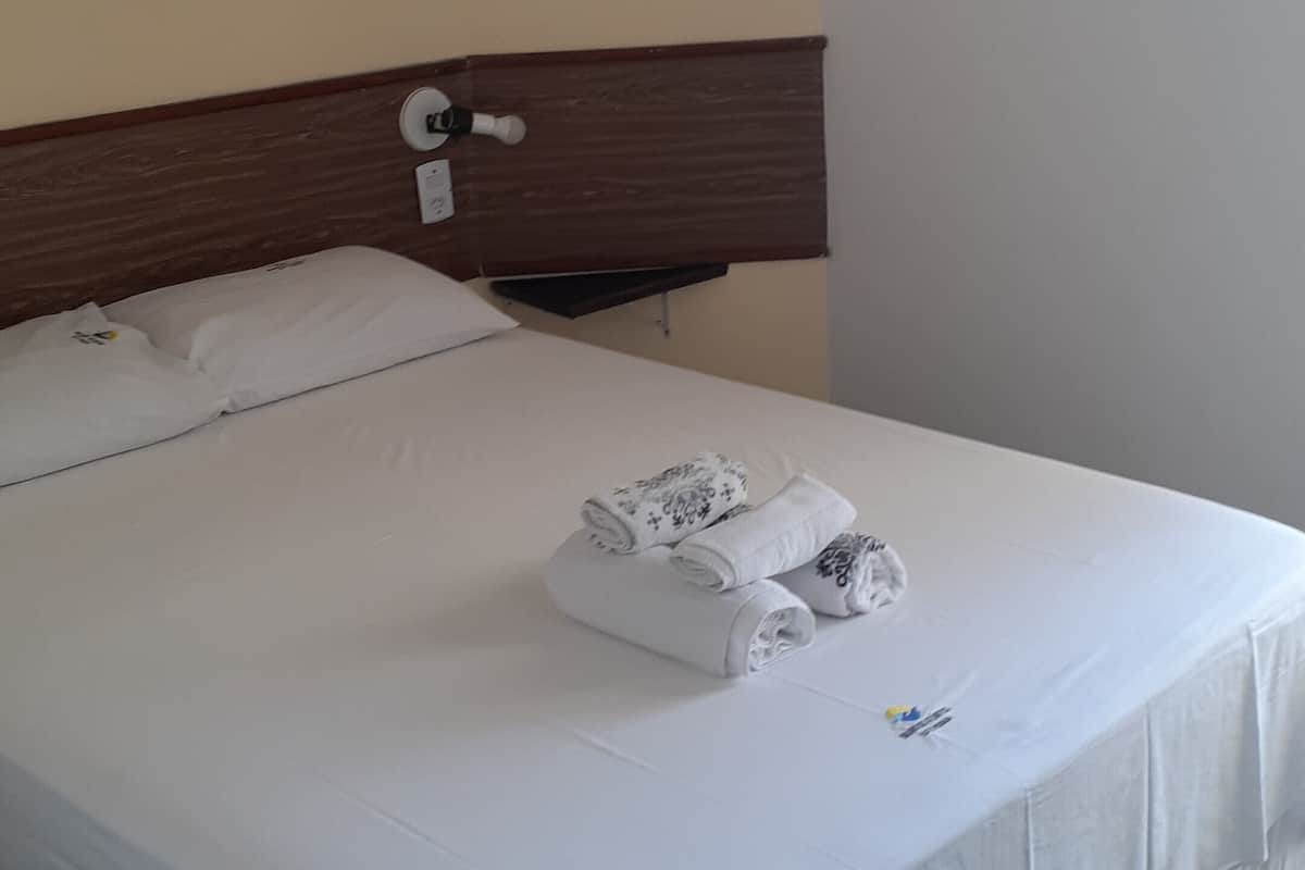 A neatly made bed with white linens and two rolled towels, a wooden headboard, and a wall-mounted light fixture.