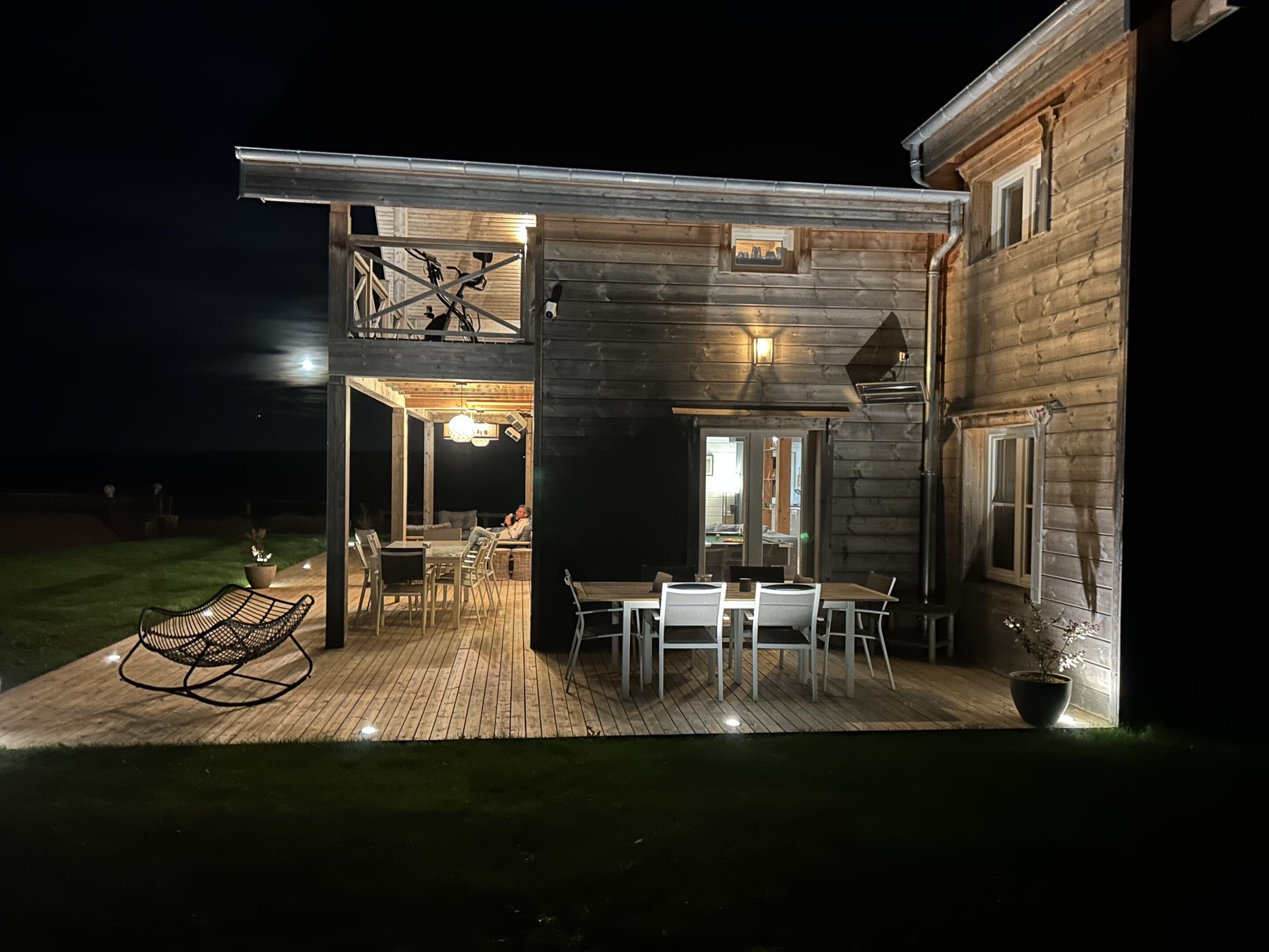 Une maison en bois à deux étages, dotée d’une terrasse, d’un espace salon extérieur et d’un escalier menant à l’étage supérieur.
