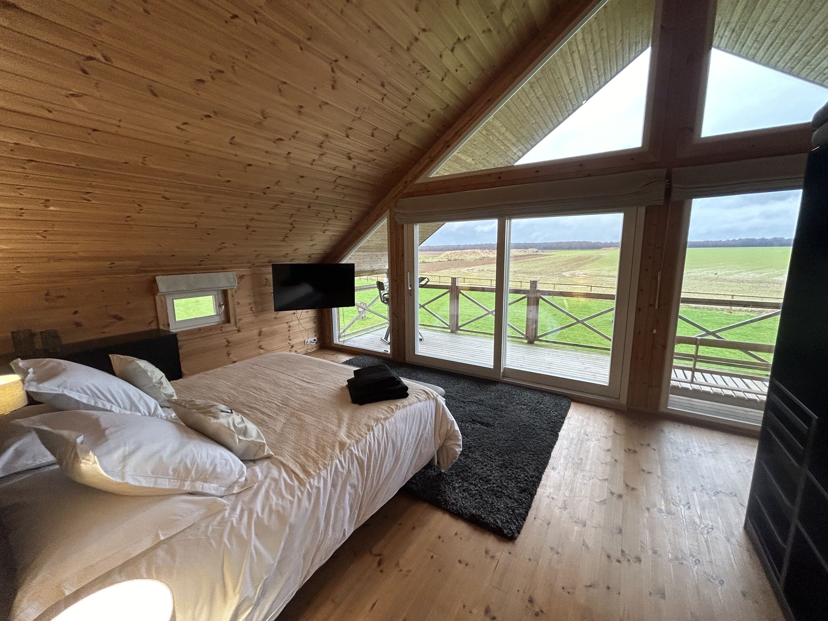 Un dormitorio de cabaña de madera con cama, almohadas, un televisor de pantalla plana y un ventanal con vistas a un campo cubierto de hierba.