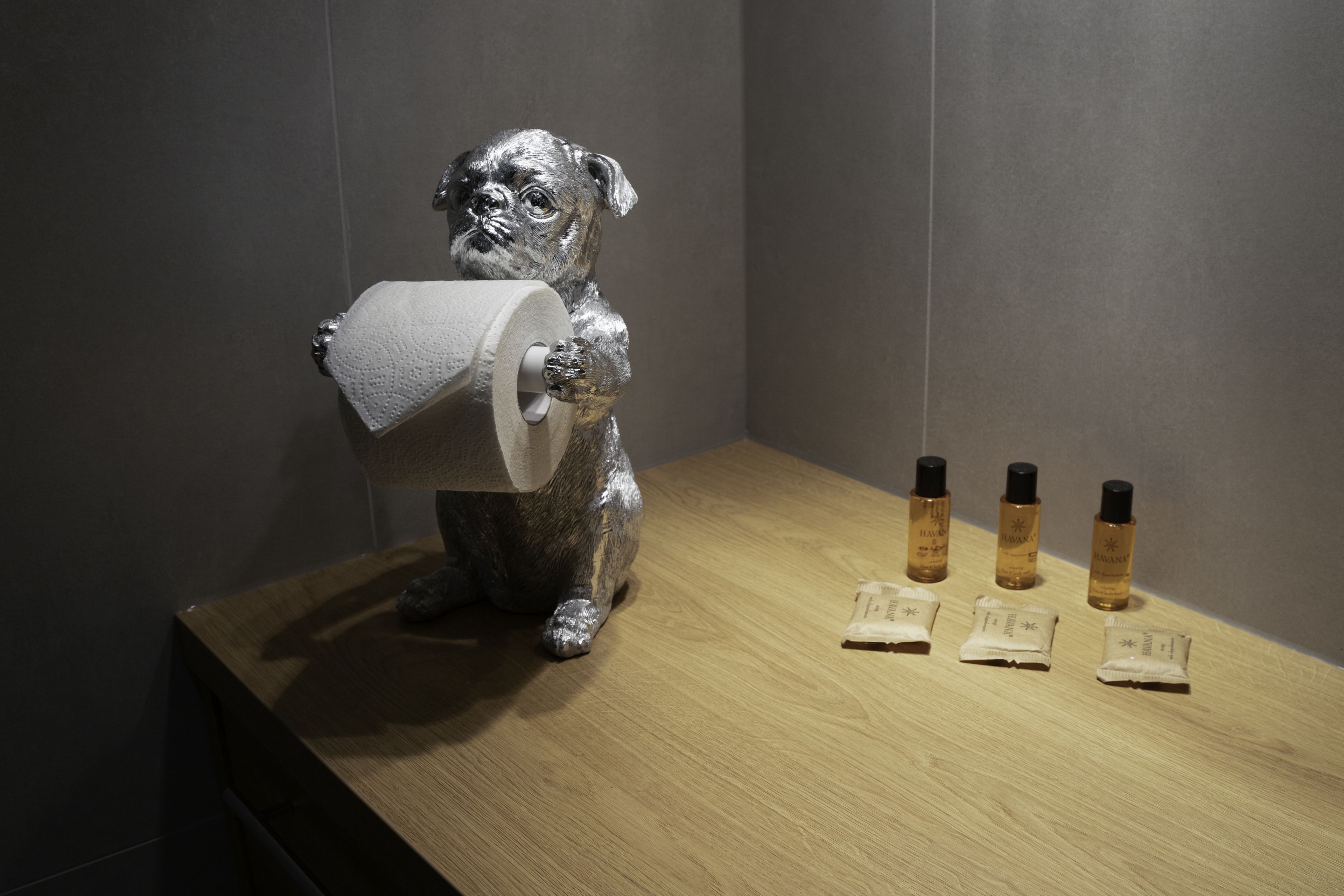 A bathroom with a wooden vanity, a silver statue of a dog holding a roll of toilet paper, and three small bottles of lotion.