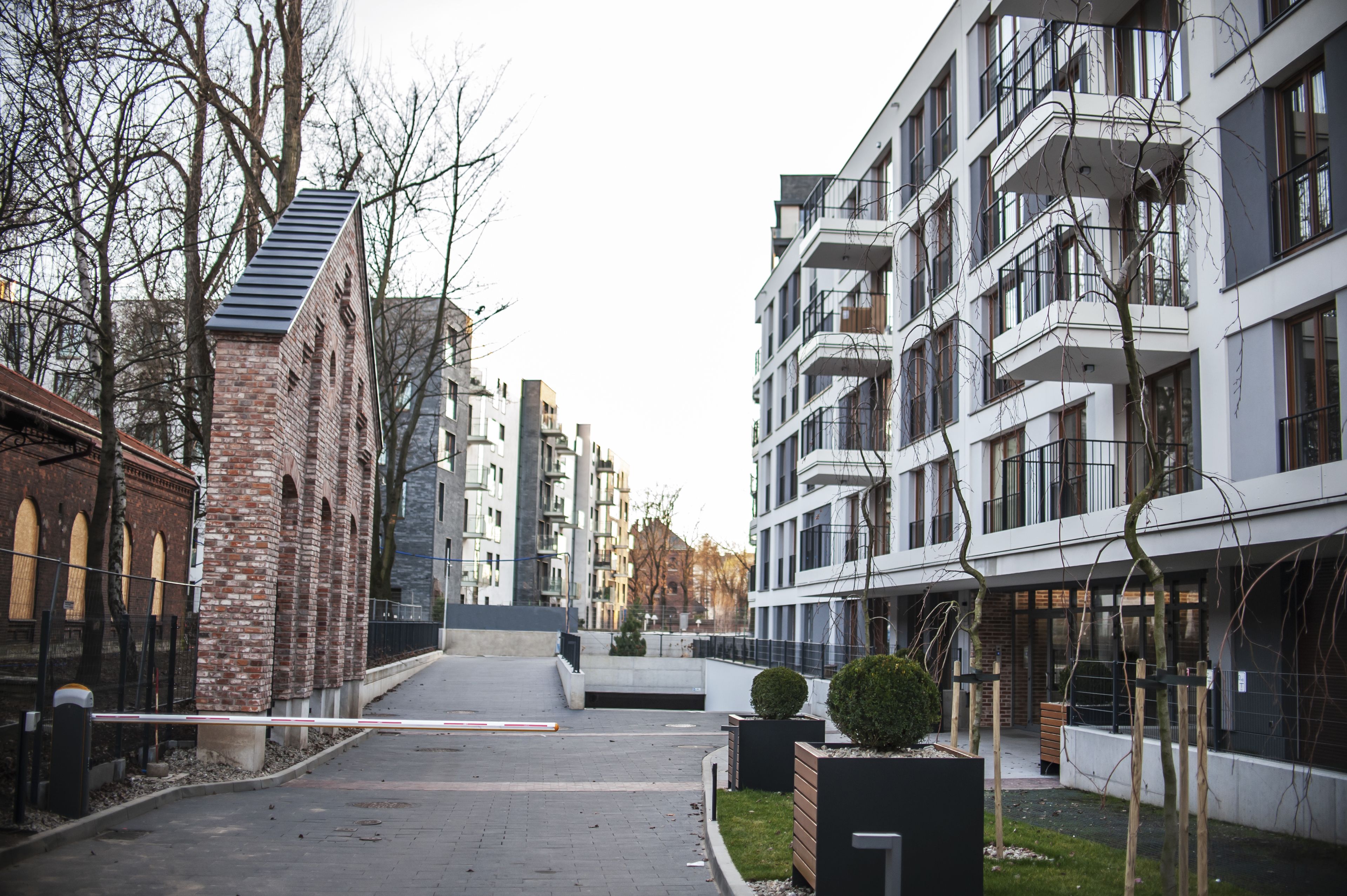 A modern urban street with multi-story buildings, a brick structure, and a gated entrance.