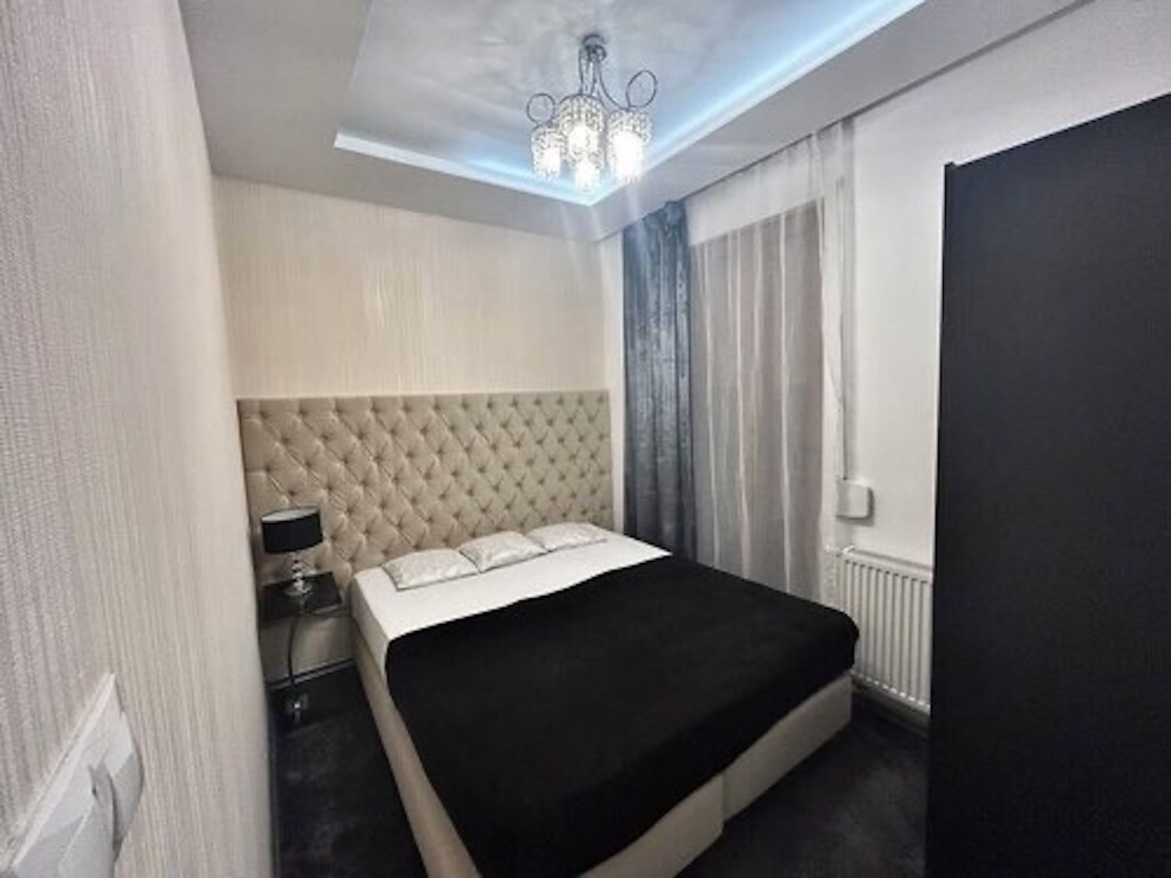 Un dormitorio moderno con cabecera capitonada, colcha negra, mesita de noche con lámpara y radiador.