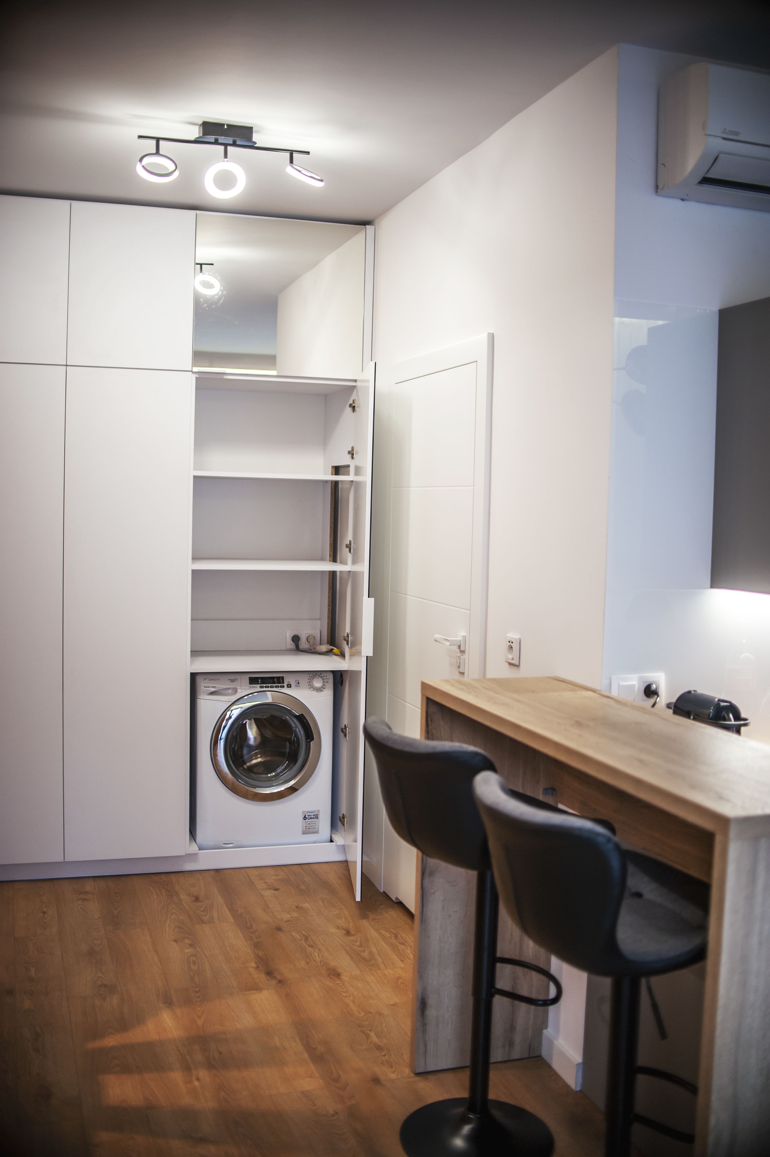A compact kitchen with a built-in washer, wooden bar stools, and a white cabinet with shelves.