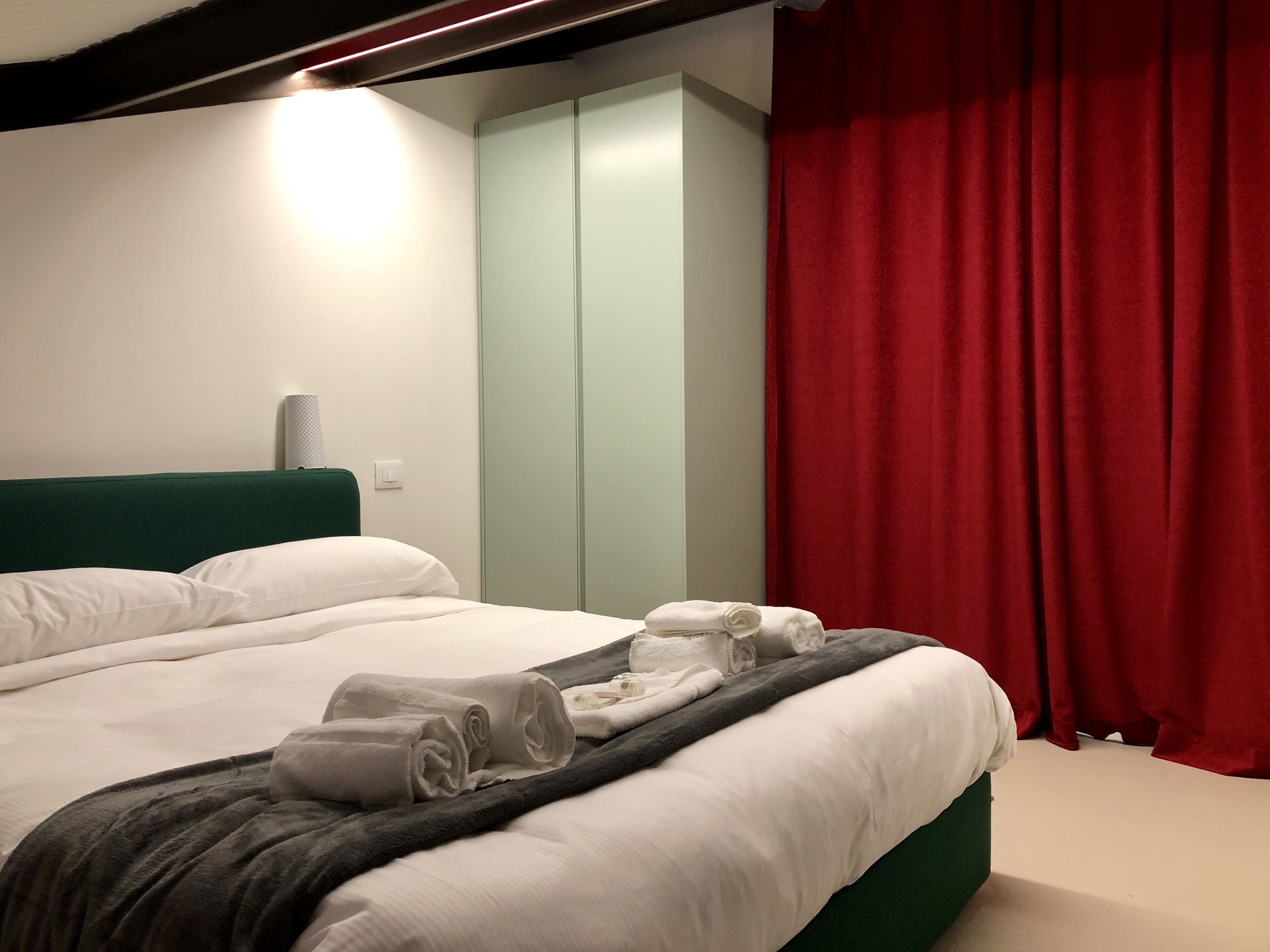 A neatly made bed with white linens and a grey blanket, a green headboard, and a red curtain.