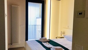 A hotel room with a bed, a window with blinds, a radiator, a nightstand with a lamp, and towels on the bed. - Rivadige Suite (Verona)