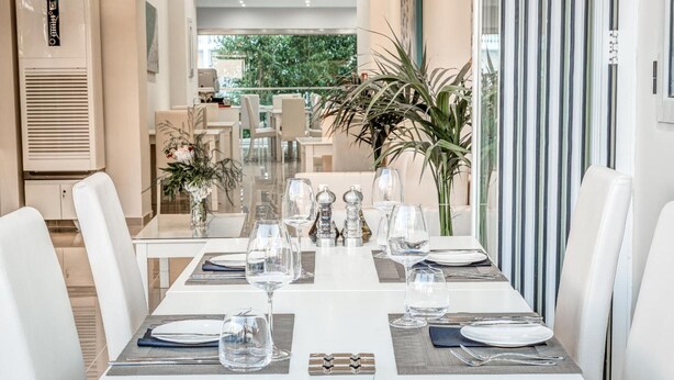 A dining area with a long table set for a meal, surrounded by white chairs, with a view of greenery outside. - Palotel Design Gouvia (Corfu)