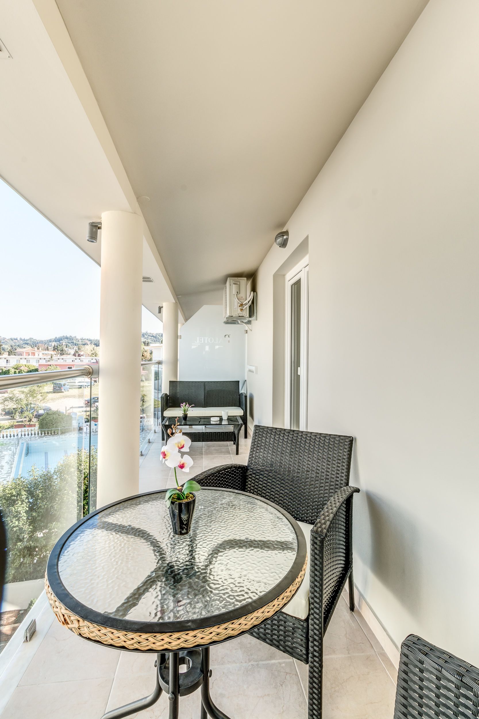 En balkong med ett runt glasbord, flätade stolar och utsikt över en pool och byggnader.