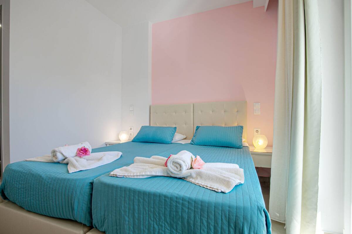 A hotel room with two beds, each with teal bedding and pillows, a tufted headboard, bedside lamps, and a pink accent wall.