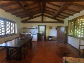 Ein geräumiges Zimmer mit hoher Decke, einem Esstisch, Stühlen, einer Küchenzeile und einem großen Fenster mit Jalousien.