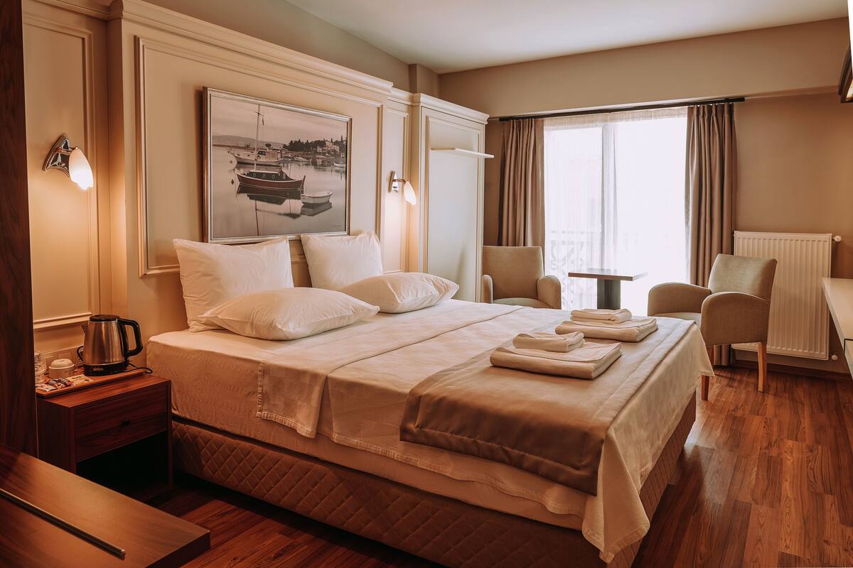A hotel room with a large bed, a desk, a chair, a radiator, and a window with curtains.