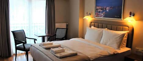 Habitación de hotel con una cama grande, dos sillas, una mesita y vistas a un paisaje urbano nocturno.