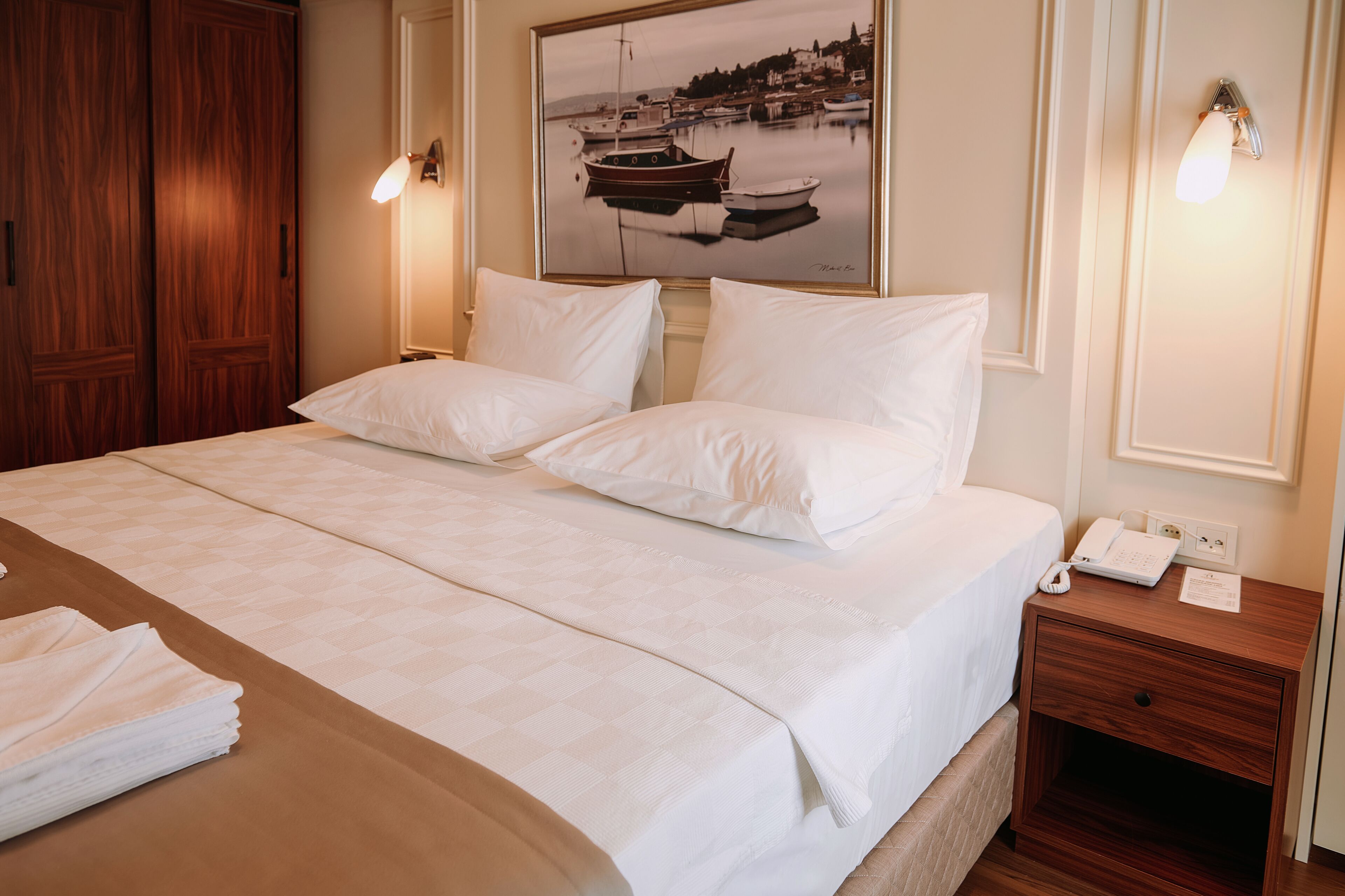 A hotel room with a large bed, a wooden nightstand, a framed picture of boats, and a wall-mounted light fixture.