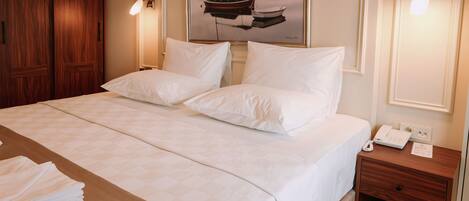 Una habitación de hotel con una cama grande, una mesita de noche de madera, un cuadro enmarcado con barcos y una lámpara de pared.