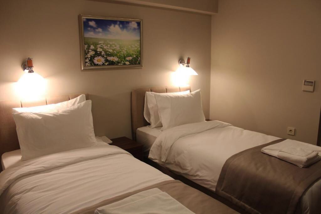 Une chambre d’hôtel avec deux lits, chacun agrémenté de draps blancs et d’un plaid marron. De chaque côté des lits, on trouve des lampes fixées au mur, et au-dessus de la tête de lit, un tableau représentant un champ de fleurs.