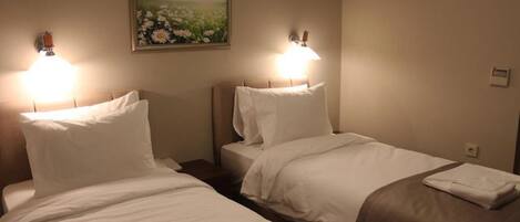 Habitación de hotel con dos camas, cada una con ropa blanca y una manta marrón. Hay lámparas fijadas a la pared a cada lado de las camas, y un cuadro de un campo con flores sobre el cabecero.