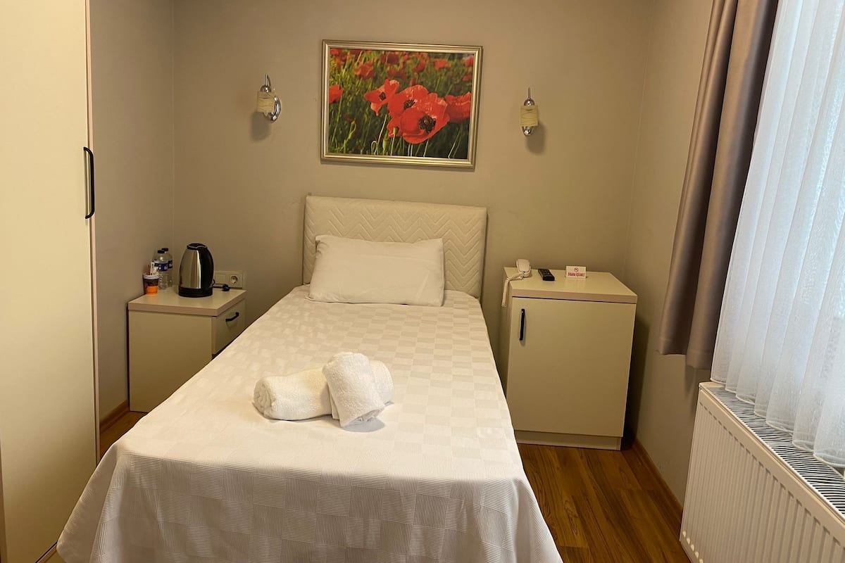 A neatly made bed with two towels, a nightstand with a coffee maker, a small cabinet, and a radiator.