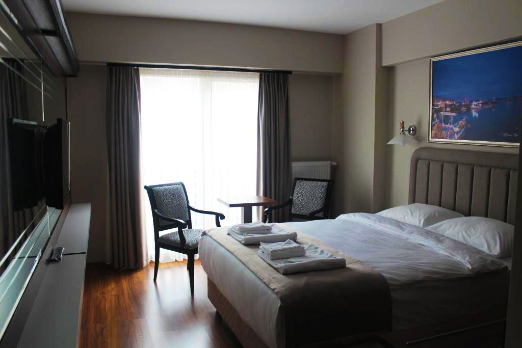 A hotel room with a bed, two chairs, a small table, a window with curtains, and a painting on the wall.