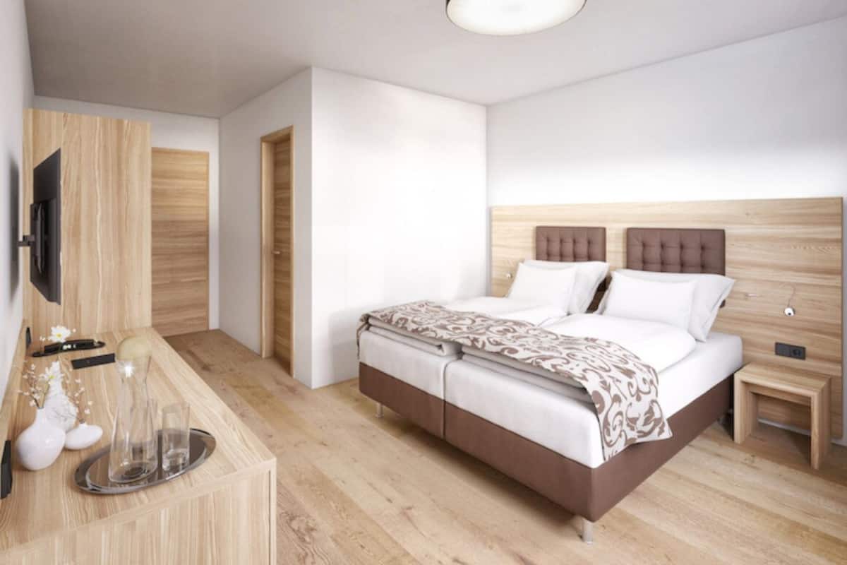 A modern hotel room with a wooden floor, a bed with a headboard, a bedside table, a TV mounted on the wall, and a small table with a vase and glasses.